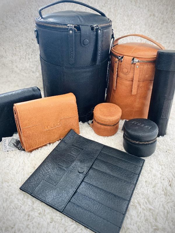 A collection of black and tan leather travel accessories, including cylindrical cases, wallets, pouches, and small zippered containers, displayed on a soft, textured surface.
