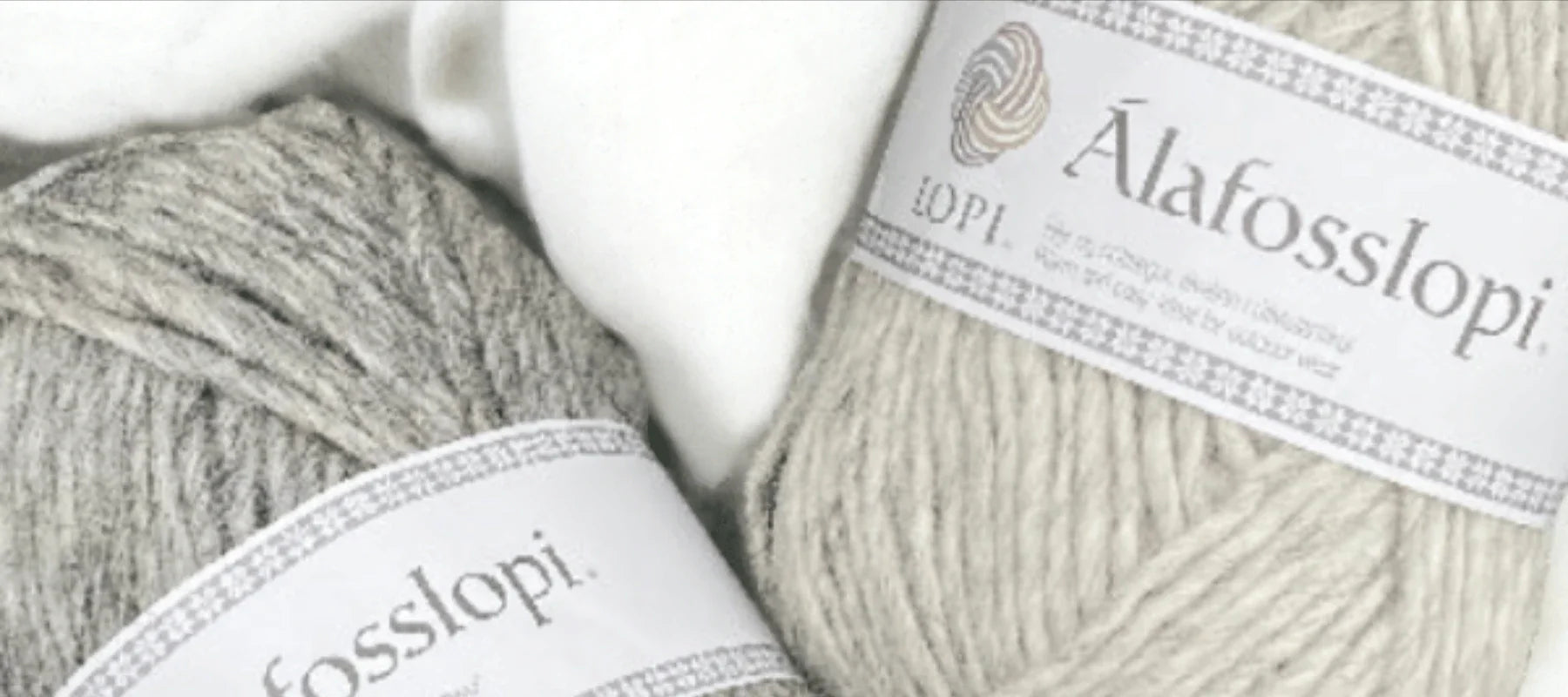 Two skeins of Álafosslopi wool yarn, one light gray and one beige, with white labels featuring Icelandic text, lie on a soft white surface.