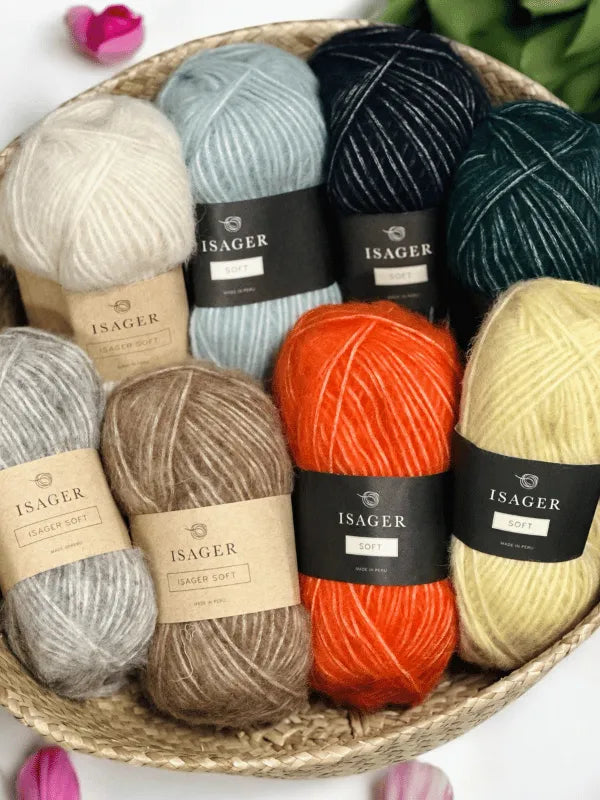 A woven basket holds eight skeins of Isager Soft in white, light blue, dark green, grey, taupe, brown, bright orange, and yellow. A few flowers peek out beside this colorful display of yarn.