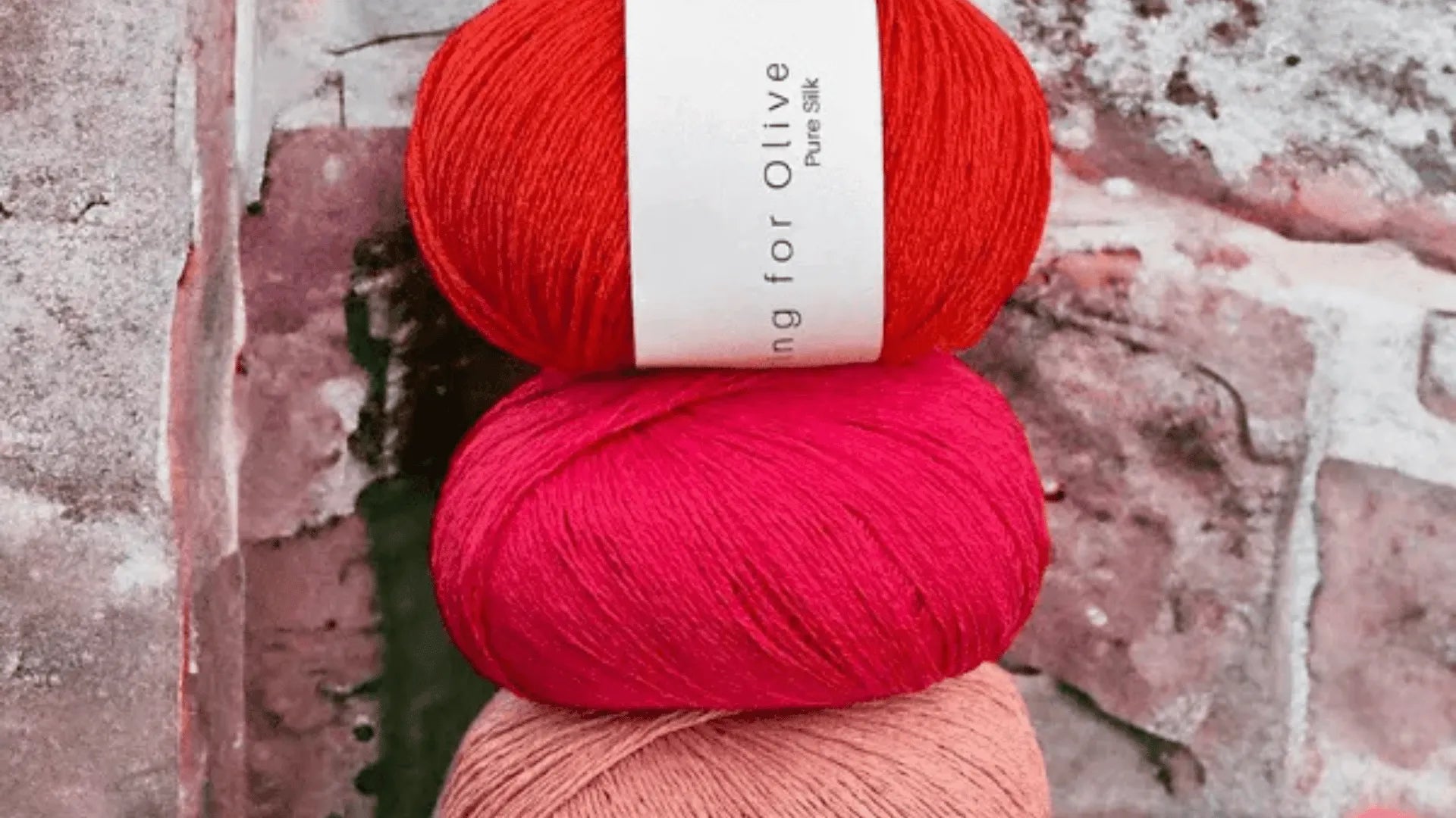 Three balls of yarn in red, pink, and peach are stacked vertically in front of a weathered, textured brick wall. The top yarn has a white label wrapped around it.