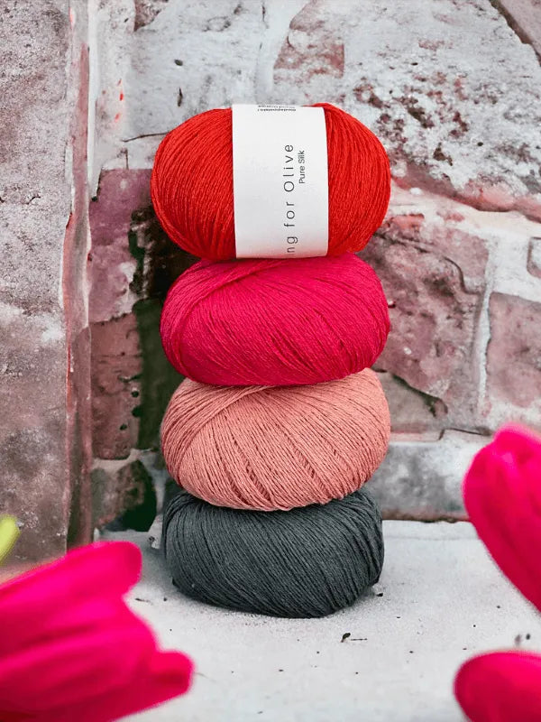 Four balls of Knitting for Olive Pure Silk yarn are stacked vertically against a stone wall in bright red, magenta, light pink, and dark gray. Blurred red flowers appear in the foreground.