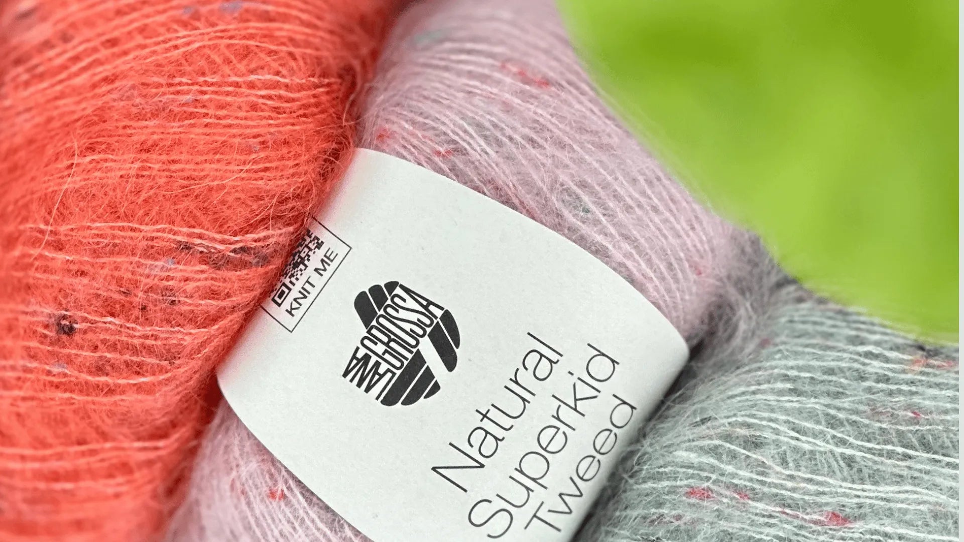 Close-up of two skeins of yarn, one coral and one pale gray with a hint of lavender, with a label reading “Natural Superkid Tweed.” The background is blurred green.
