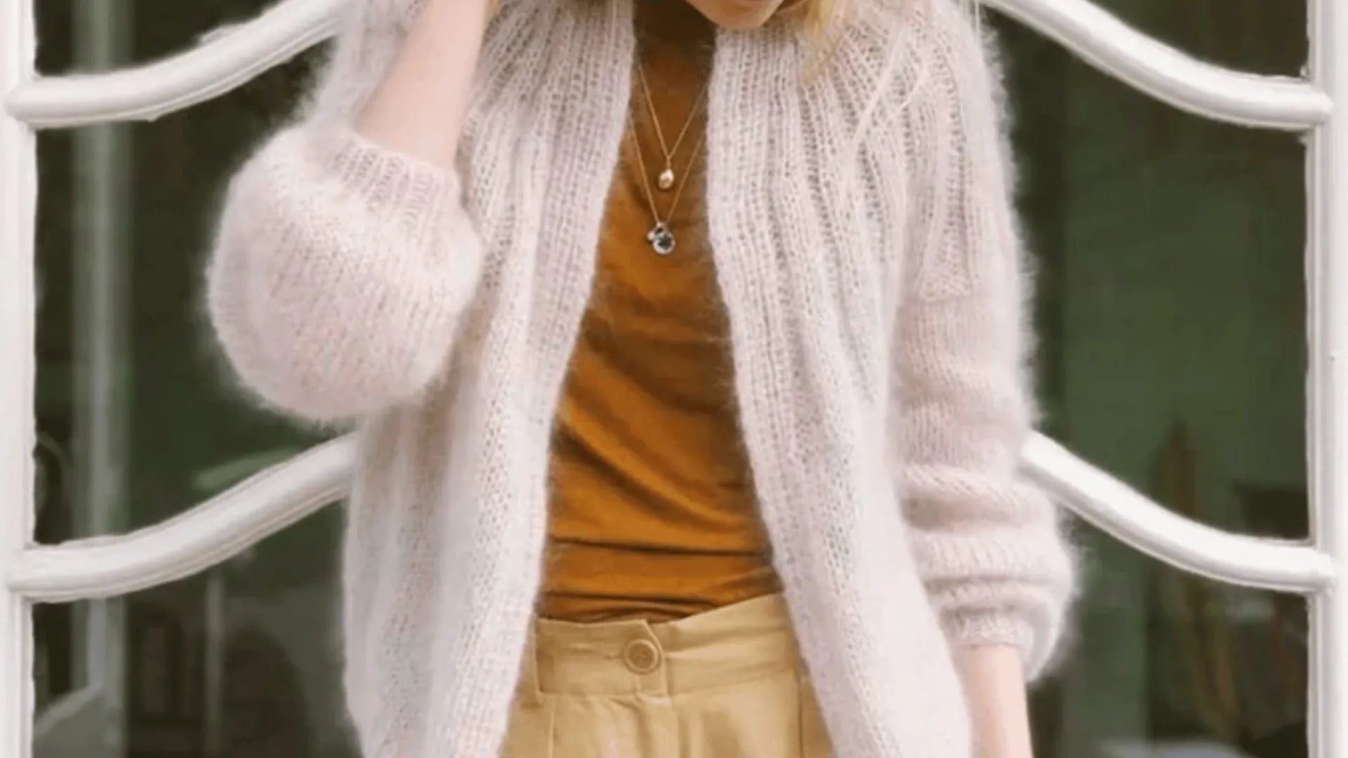 A person wearing a fuzzy cream cardigan over a brown top and beige high-waisted pants stands outside, with one hand touching their head. Only the torso and part of the head are visible.