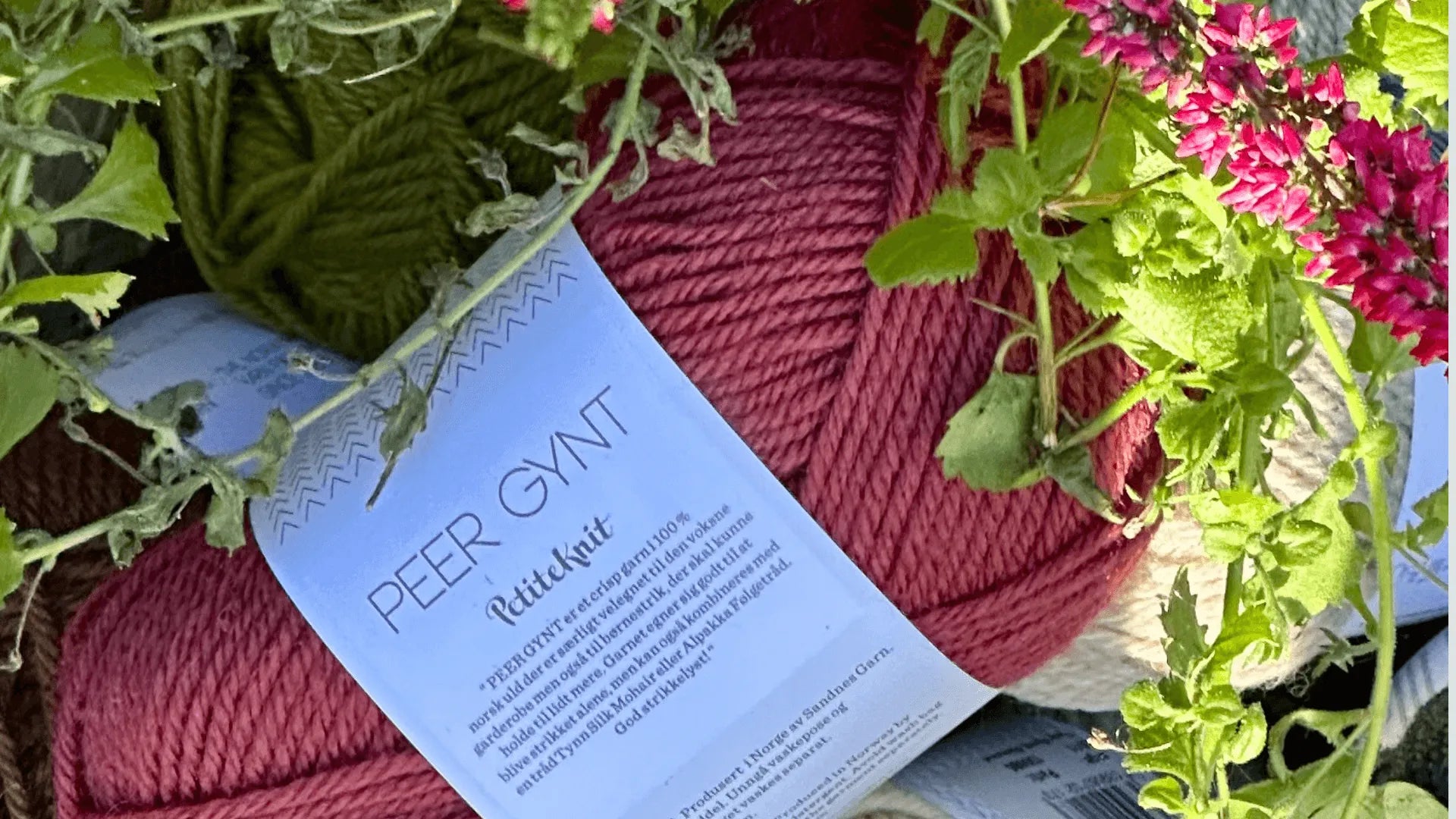 A close-up of a skein of Peer Gynt Rødtet knit yarn in rose pink, surrounded by green leaves and clusters of small bright pink flowers.