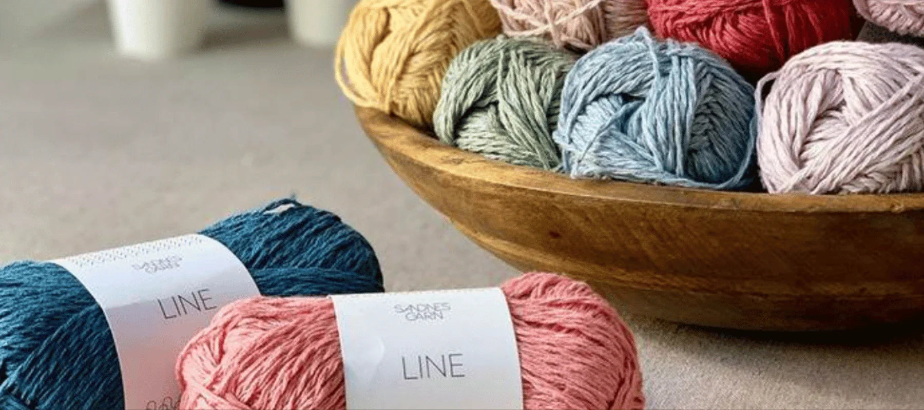 A wooden bowl holds several skeins of colorful yarn, with two skeins—one teal and one pink—labeled LINE by Sandnes Garn placed in the foreground on a beige surface.