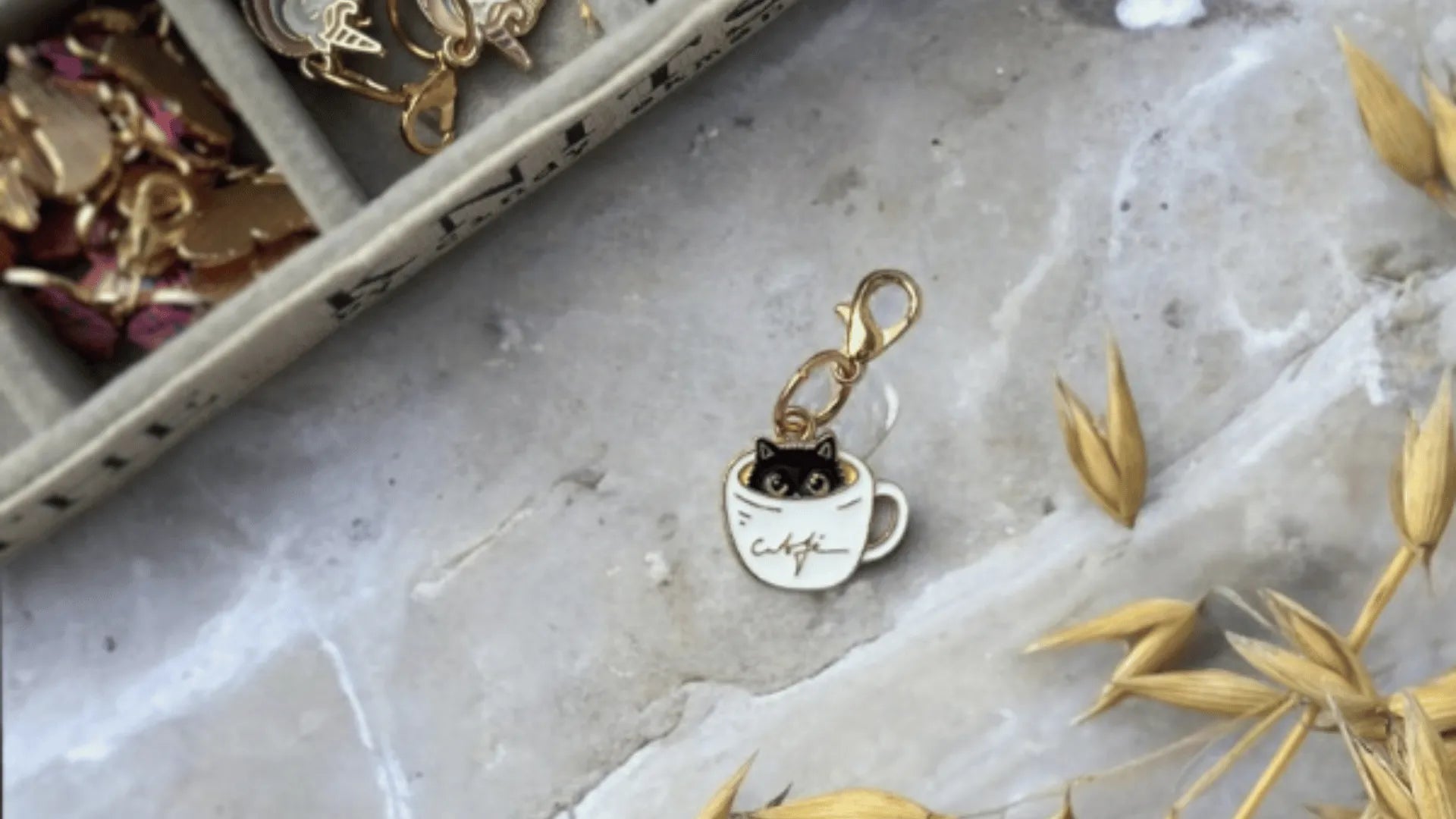 A gold keychain charm featuring a black cat sitting inside a white coffee cup with coffee written on it lies on a marble surface next to gold jewelry and dried golden leaves.