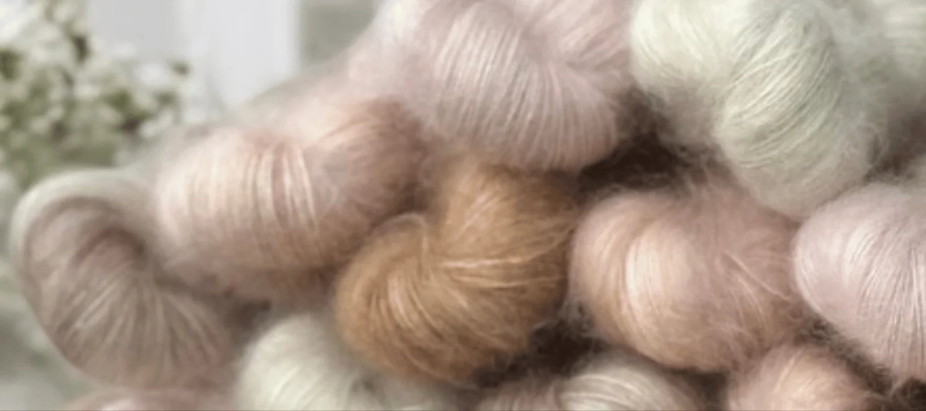A close-up of several skeins of soft, fluffy yarn in pastel shades of cream, blush pink, and light brown, stacked together in a cozy arrangement.