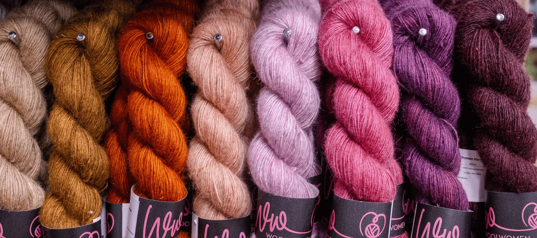 A row of soft, fluffy yarn skeins in various shades of brown, orange, pink, purple, and burgundy are neatly arranged side by side with black labels at the bottom.