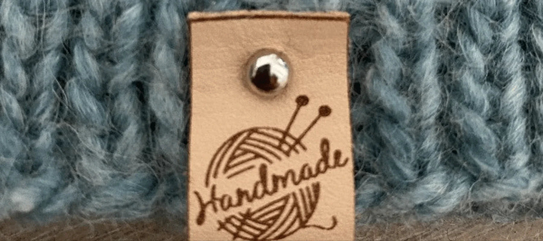 A close-up of a blue knitted fabric with a beige leather tag attached by a metal rivet. The tag is stamped with a ball of yarn, knitting needles, and the word Handmade.