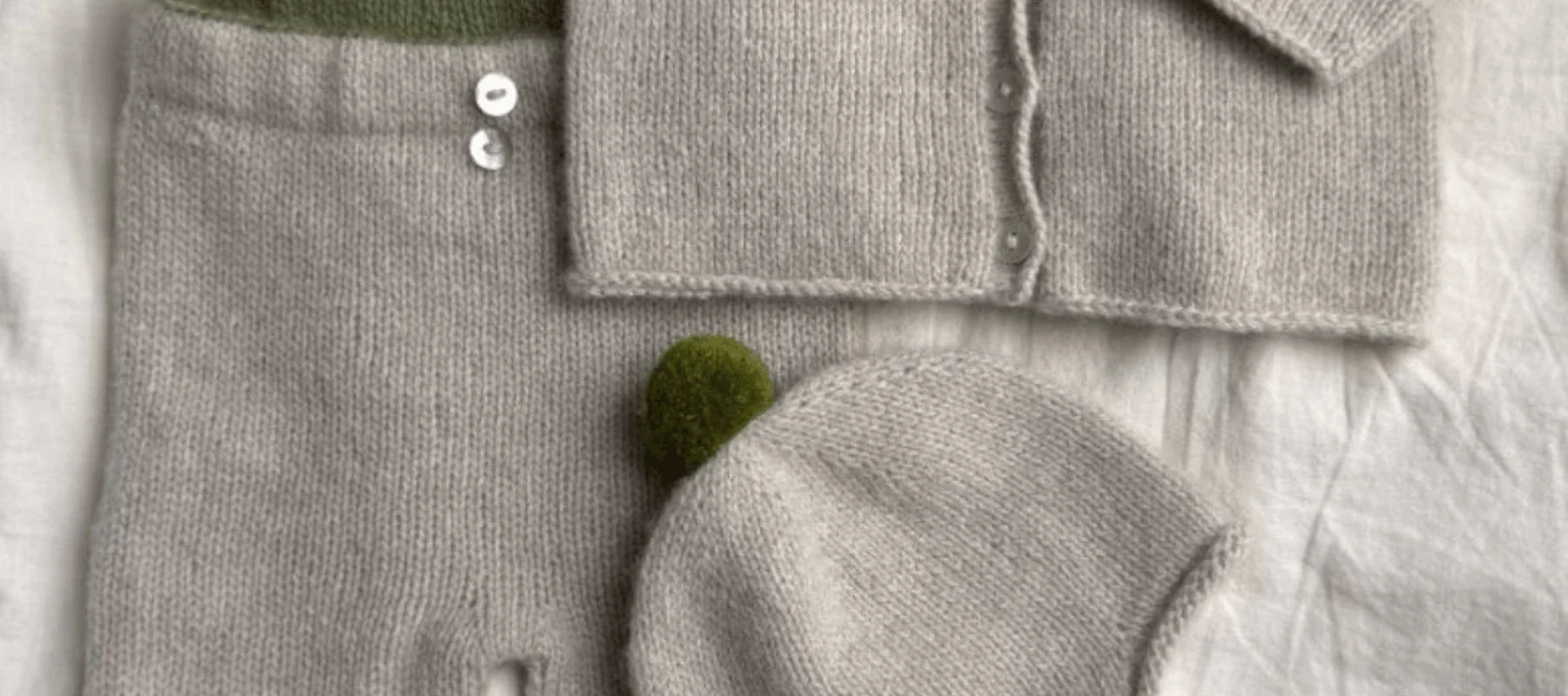 A soft, light gray knitted baby outfit with pants, a buttoned cardigan, and a hat featuring a small green pom-pom, all laid out on a white fabric background.
