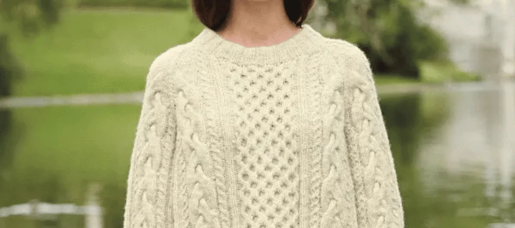 A person wearing a cream-colored, cable-knit sweater stands outdoors in front of a pond and greenery.