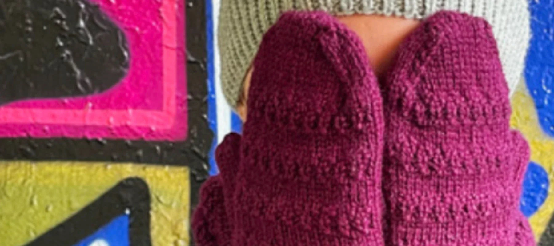 A person wearing a gray knit beanie holds up two maroon knitted mittens in front of a colorful, abstract painted wall.