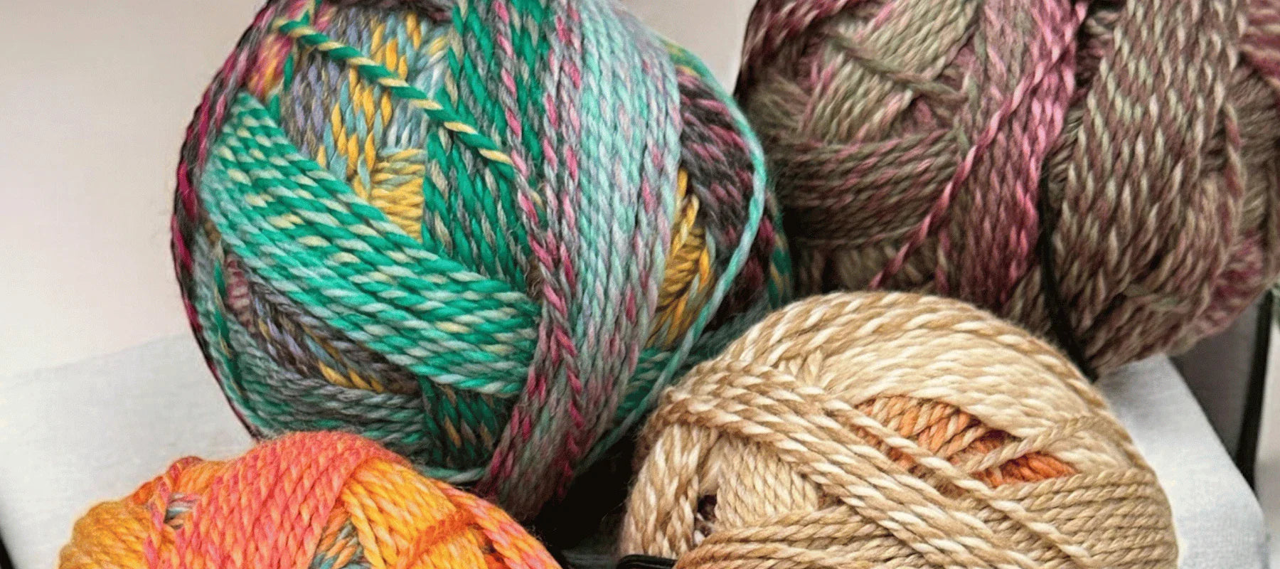 Four balls of colorful yarn, each with a different blend of colors including teal, yellow, orange, pink, beige, and brown, are displayed on a surface against a neutral background.