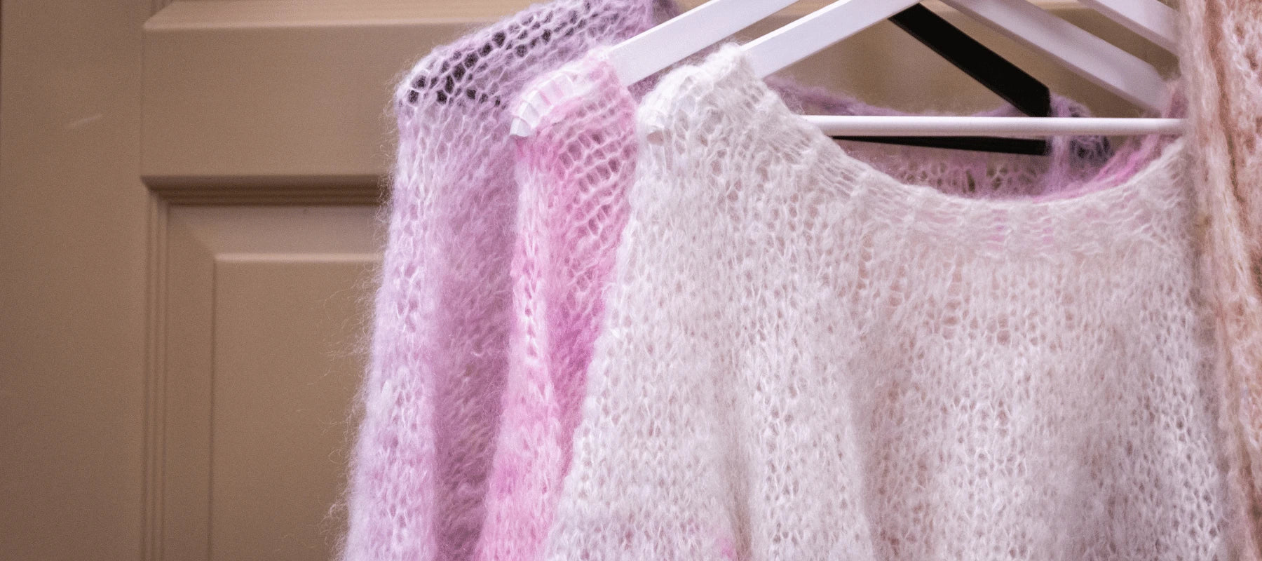 Three soft, fluffy knitted sweaters in pastel pink and white hang on white hangers against a beige door background. The texture appears cozy and delicate.