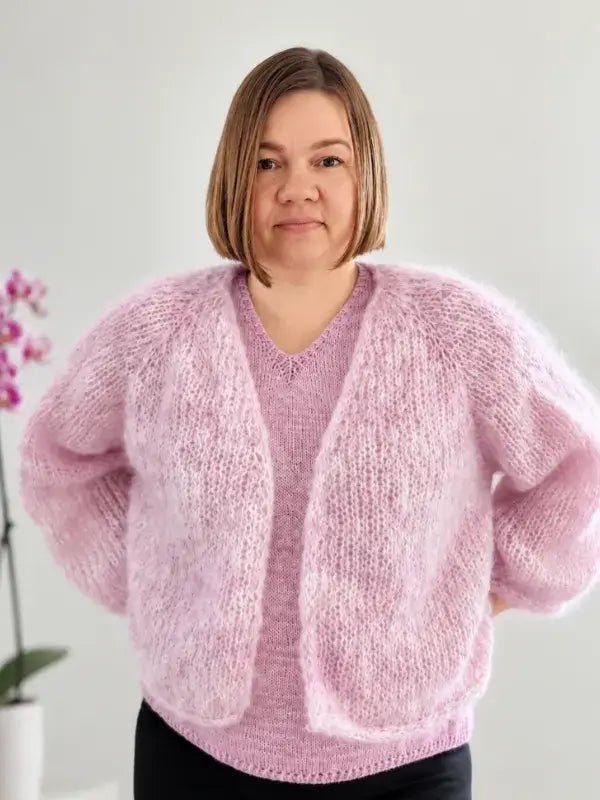 A woman with short brown hair, wearing a light purple knit sweater and the Fluff neuletakki ohje, stands indoors with her hands behind her back. A pink orchid is visible in the background.