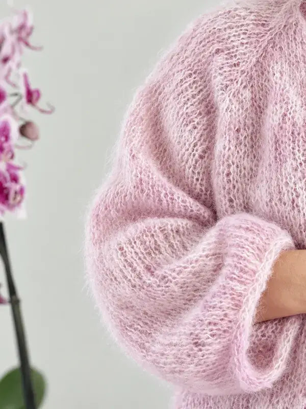 A person wearing a soft, fluffy light pink Fluff neuletakki ohje + WoolWomen Fluff Mohair stands next to a pink orchid flower. Only part of the arm and torso are visible, highlighting the sweater’s mohair lanka texture and delicate color.