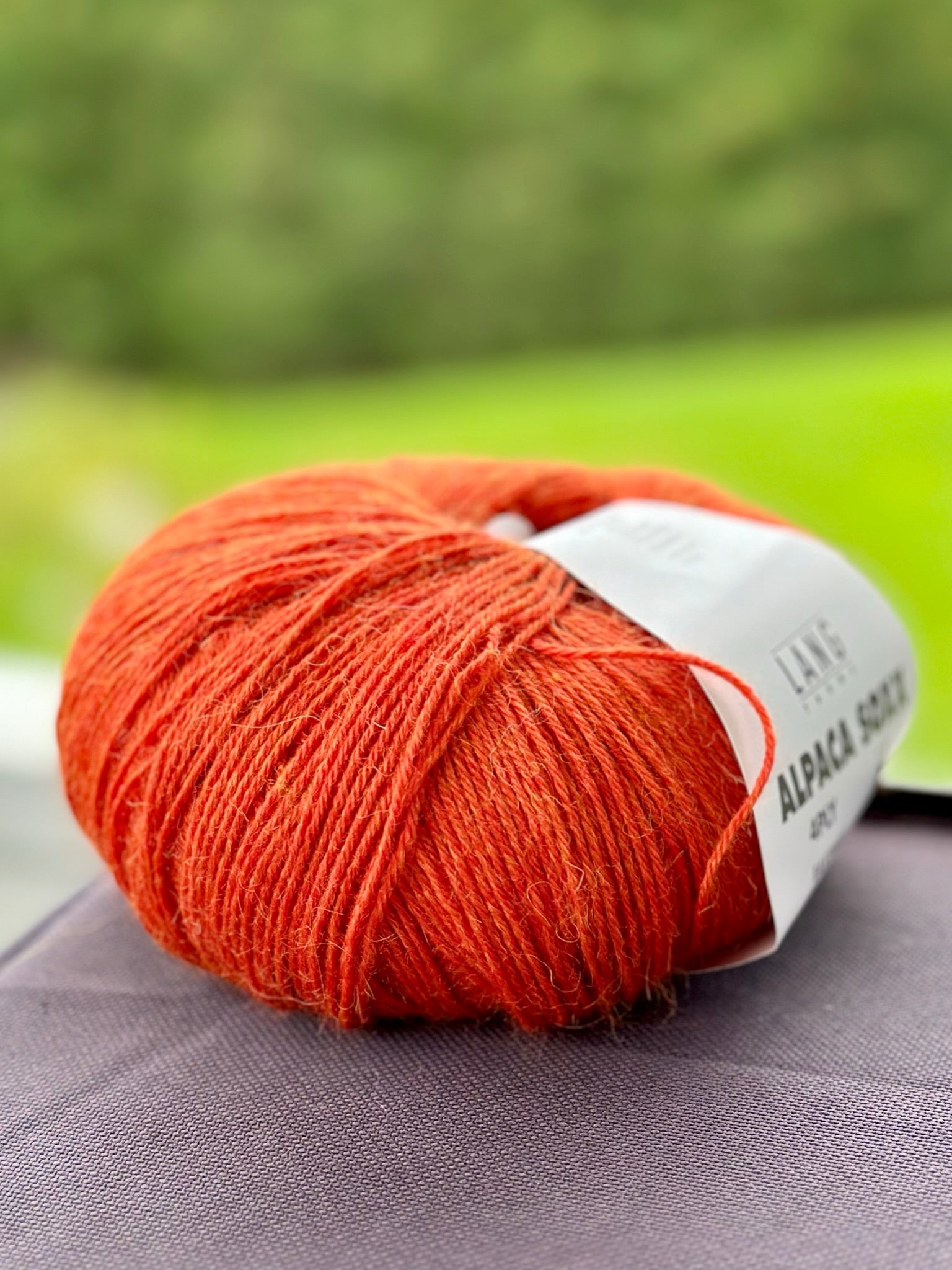 A ball of bright orange Lang Alpaca Soxx 4ply yarn sits on a dark surface, with Mittums lapasten ohje (PDF) shown in the background and a blurred green outdoor scene behind.