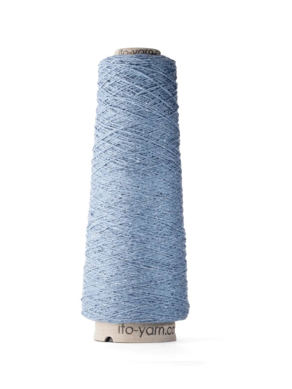 A tall cone of light blue Ito Kinu stands upright, its silk thread tightly wound around a white paper core marked ito-yarn.com—ideal for kesäneuleisiin.