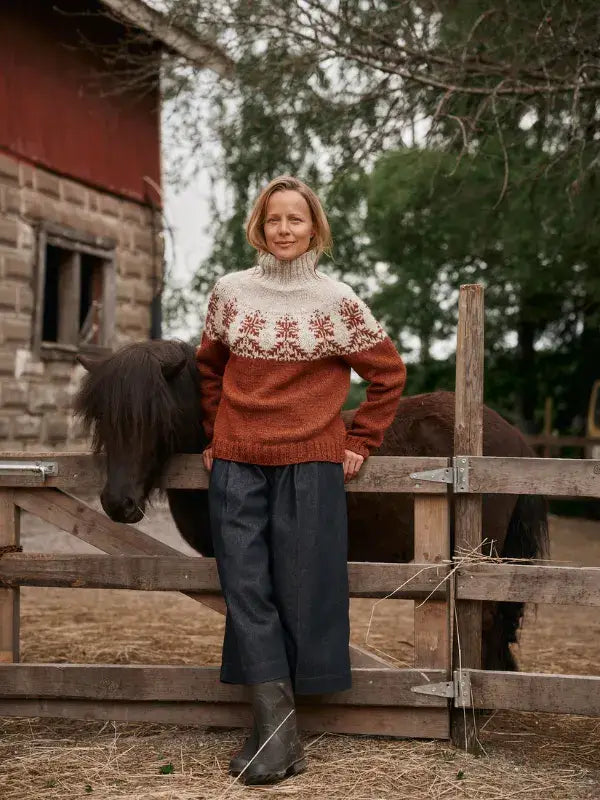 A woman wearing a rust and cream-colored Jenna Kostet - Neulottu Kalevala III, Ennakkotilaus patterned wool sweater stands next to a dark horse by a wooden fence.
