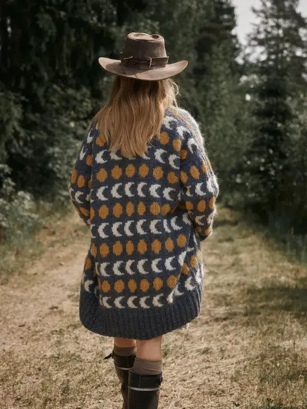 Person wearing a felt hat and a dark blue Jenna Kostet - Neulottu Kalevala III, Ennakkotilaus cardigan with a pattern of yellow dots and white crescent moons, walking along a forest path.