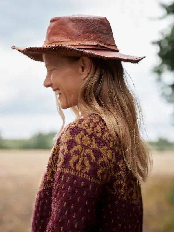 A person wearing a wide-brimmed leather hat and a patterned maroon and gold Neulottu Kalevala III knit sweater.