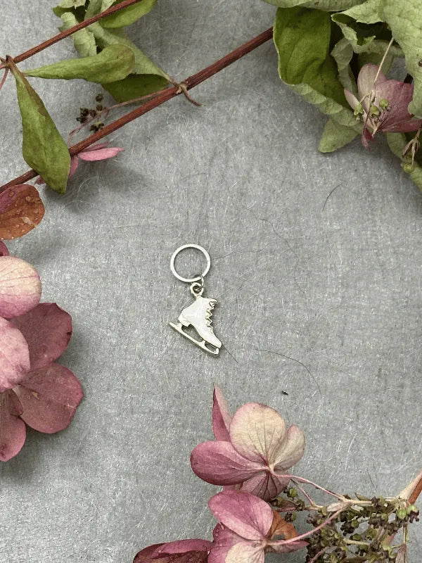 A small silver ice skate charm from KNITS by cindy ekman silmukkamerkit rests on a gray surface with pink hydrangea flowers and green leaves—ideal for neulonta lovers seeking elegant stitch markers.