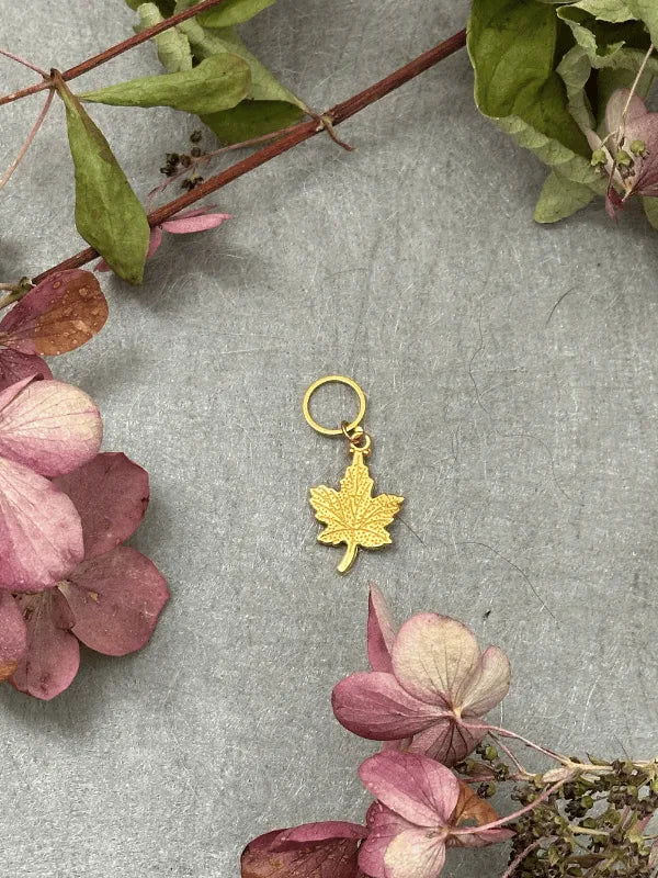 A KNITS by cindy ekman silmukkamerkit gold maple leaf charm with a small hoop lies on a gray surface, surrounded by dried pink and green leaves and flowers—perfect for neulonta projects.