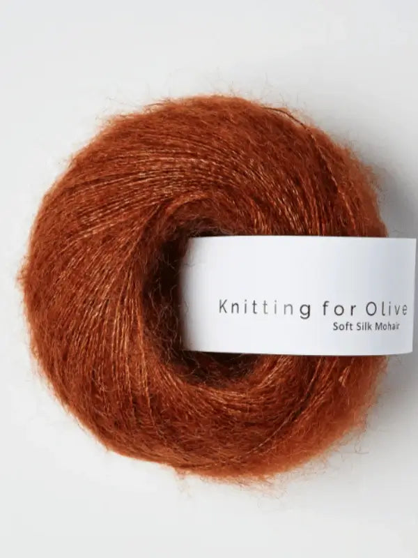 A ball of rust-colored Knitting for Olive Soft Silk Mohair yarn, featuring a white label, is displayed against a plain light background. Made from luxurious kid mohair and silkki lanka, this yarn meets the Responsible Mohair Standard for quality and ethical sourcing.