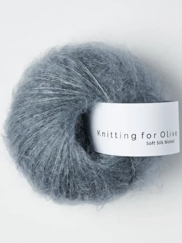 A ball of fluffy blue-grey Knitting for Olive Soft Silk Mohair yarn, featuring a white label, made from vastuullinen silk and kid mohair fibers, is shown against a light background.