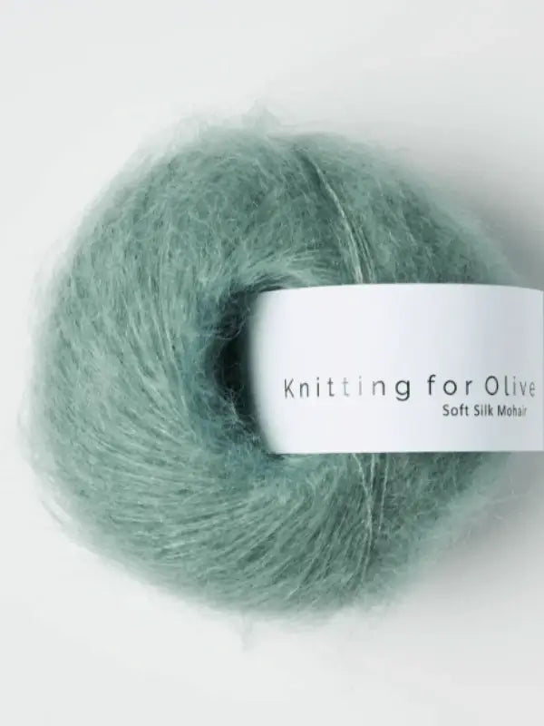 A ball of soft, light green Knitting for Olive Soft Silk Mohair yarn with a white label sits against a plain light background; this kid mohair-silk yarn is responsibly sourced according to the Responsible Mohair Standard.