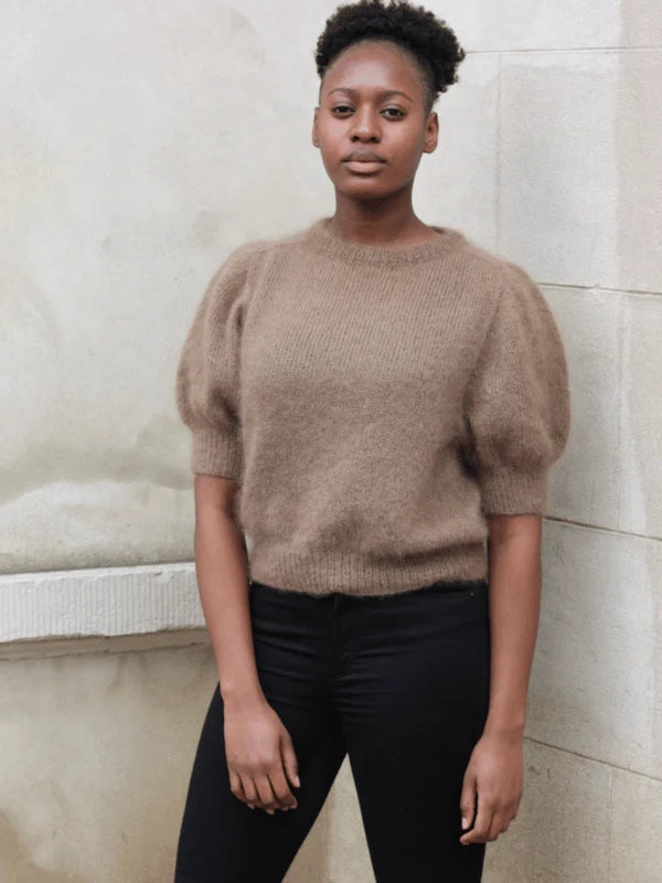 A woman with short, natural hair stands by a light wall, wearing a brown Puff Tee neuleohje fuzzy knit sweater with puffed sleeves and black pants, her calm gaze highlighting the stylish mohair top.