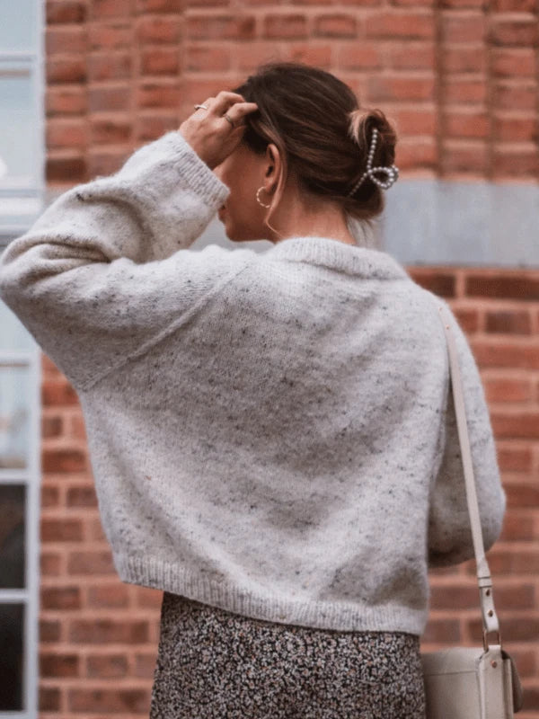 A woman with brown hair clipped up, wearing the KutovaKika Cinema Paita sweater knitted with Sandnes Garn Sunday yarn and a patterned skirt, stands outdoors in front of a brick wall, facing away from the camera and touching her hair.