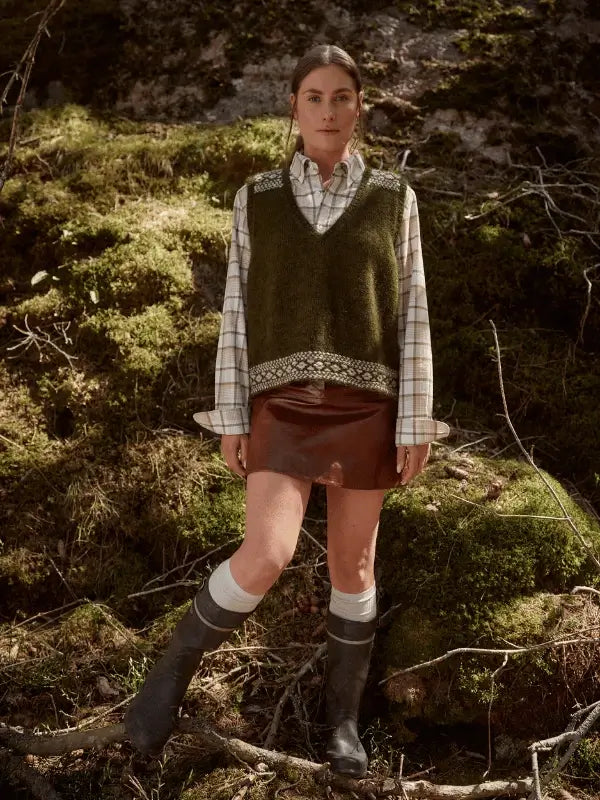 A woman stands on mossy ground in a green knitted vest—perhaps crafted from Laine Juhlanumero - ENNAKKO patterns—layered over a plaid shirt, brown skirt, white knee-high socks, and black boots among sunlit trees, channeling Nordic style.