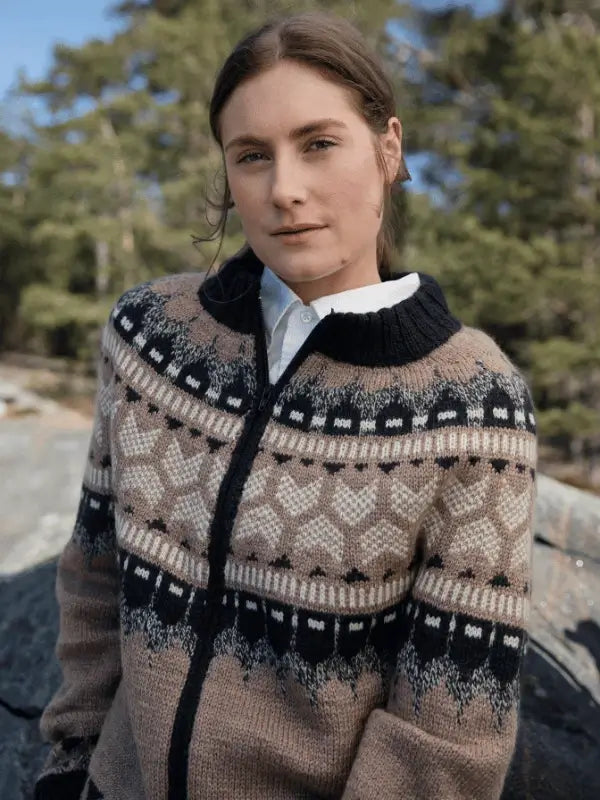 A person with brown hair, dressed in a patterned wool sweater inspired by pohjoismainen estetiikka from Laine Juhlanumero - ENNAKKO over a white collared shirt, stands outdoors among trees and rocks, looking calmly at the camera.