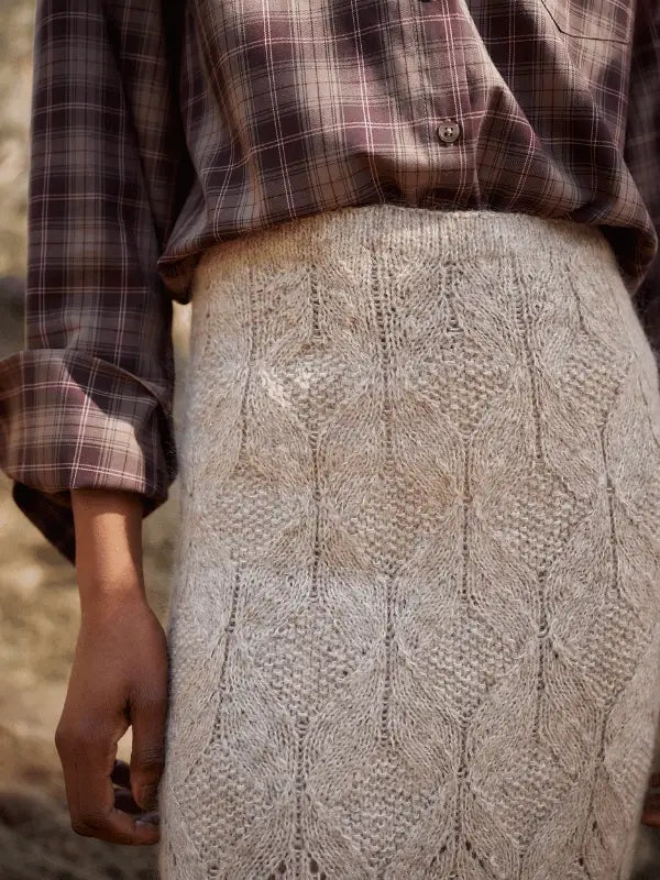 A person wears Laine Juhlanumero - ENNAKKO: a brown plaid long-sleeve shirt tucked into a textured, light beige skirt with a leafy pattern, capturing pohjoismainen estetiikka. Only the torso, arm, and top of the skirt are shown.