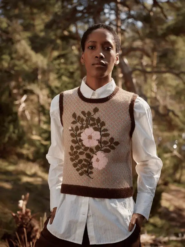 A person stands outdoors among trees, wearing Laine Juhlanumero - ENNAKKO—a white shirt and beige floral knit vest—capturing pohjoismainen estetiikka as sunlight filters through the forest in the background.