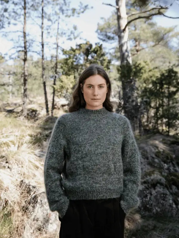 A person with long brown hair, wearing a thick gray sweater inspired by pohjoismainen estetiikka from Laine Juhlanumero - ENNAKKO and black pants, stands outdoors amid rocks, trees, and greenery.