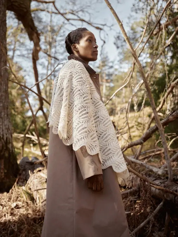 Standing outdoors among trees in a long beige coat and white lacy shawl, this scene captures the timeless Nordic aesthetic featured in Laine Juhlanumero - ENNAKKO.
