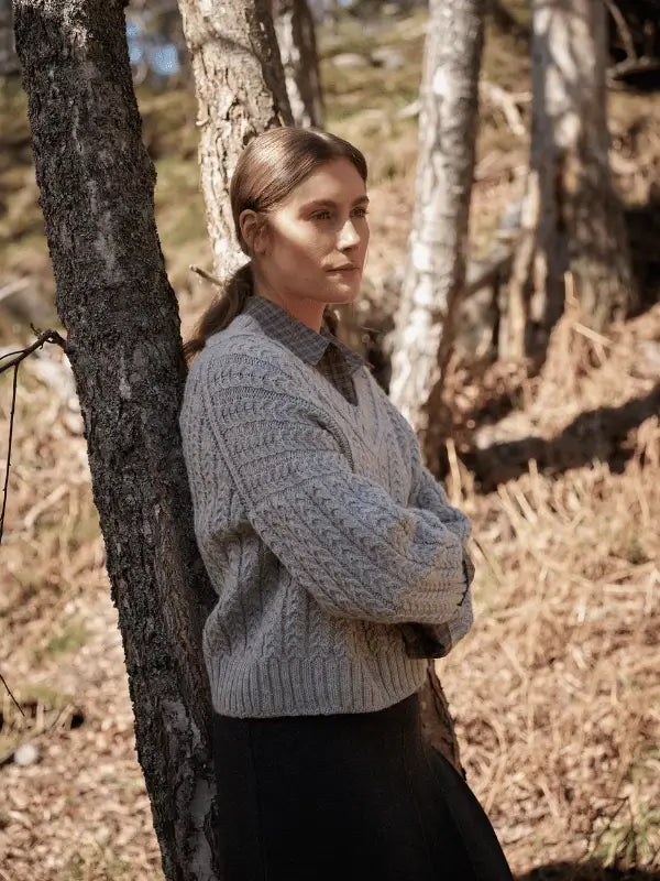 A woman with light brown hair tied back, wearing a thick gray cable-knit sweater inspired by neulekaavat from Laine Juhlanumero - ENNAKKO, stands arms crossed against a sunlit forest tree, embodying pohjoismainen estetiikka.