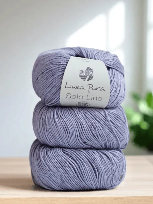 Three balls of light purple Lana Grossa Solo Lino yarn are stacked on a wooden surface, with a blurred green plant and window in the background.