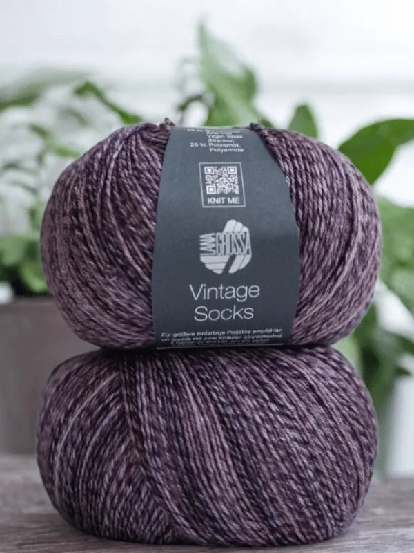 Two skeins of Lana Grossa Vintage Socks yarn are stacked on wood with a blurred green plant behind—perfect for your next project with the Lauri Fingerin sukkaohje + Lana Grossa Vintage socks set.