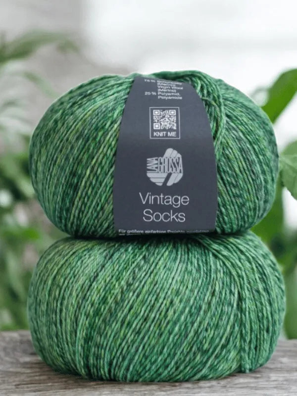 Two skeins of Lana Grossa Vintage Socks yarn from the Lauri Fingerin sukkaohje +Lana Grossa Vintage socks set are stacked on wood, with a dark gray label showing white text and a QR code. Blurred greenery appears in the background.