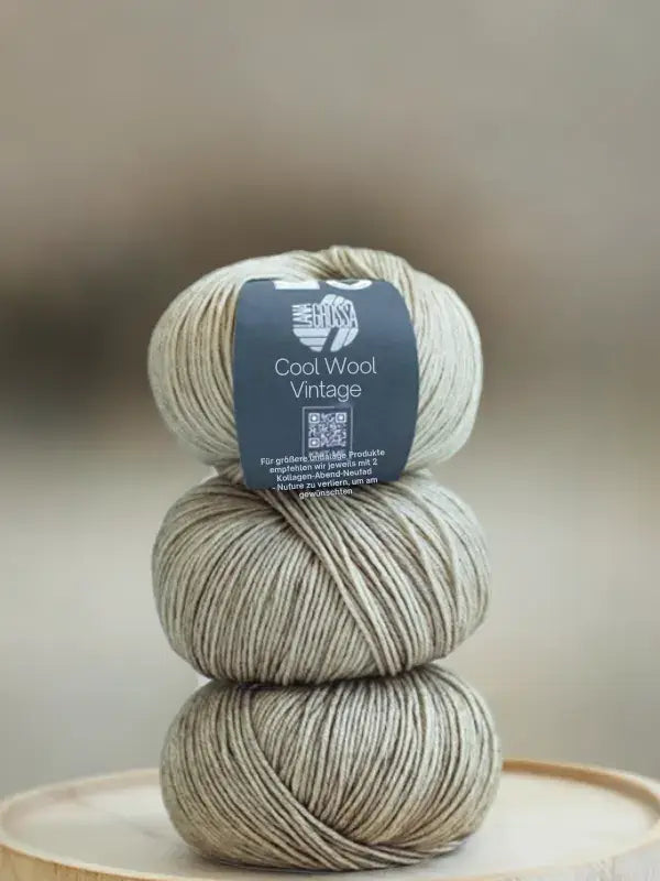 Three stacked balls of beige, heathered Lana Grossa Cool wool vintage yarn with a label around the top ball.