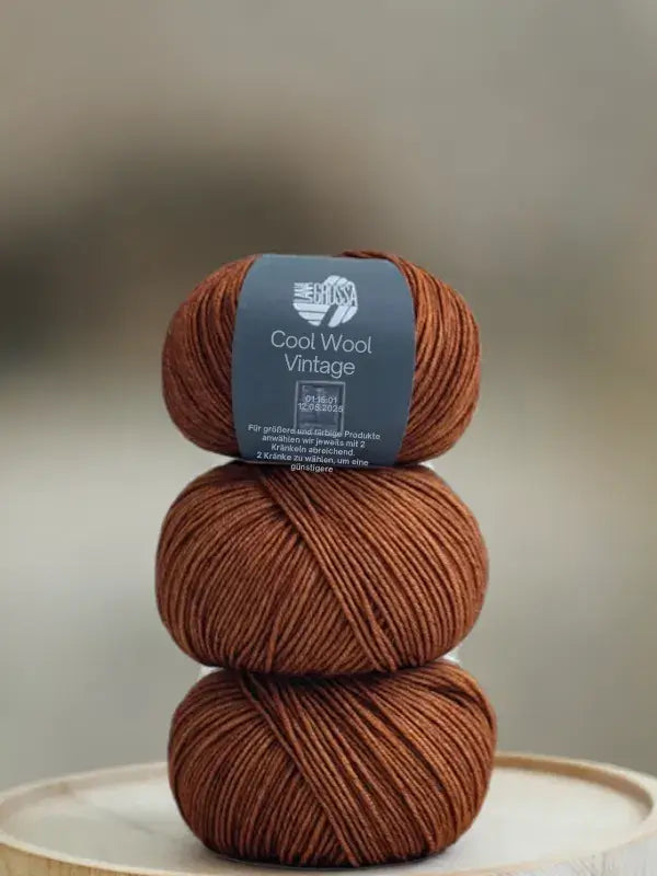 Three balls of Lana Grossa Cool wool vintage yarn in a rust color stacked vertically, with a label on the top ball featuring text in German.
