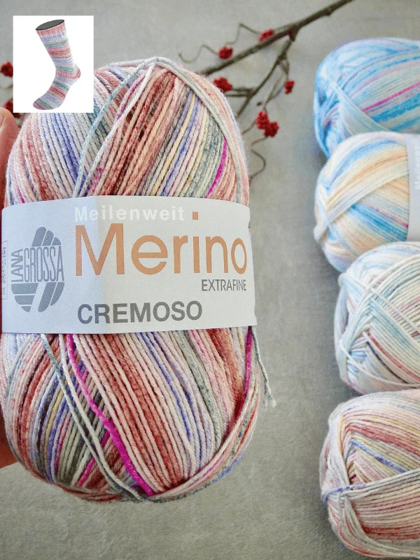 A hand holds a multicolored ball of Lana Grossa Meilenweit 100 Merino Extrafine Cremoso yarn; three similar machine-washable balls and a branch with red berries are in the background, with an inset showing a sock made from the yarn.