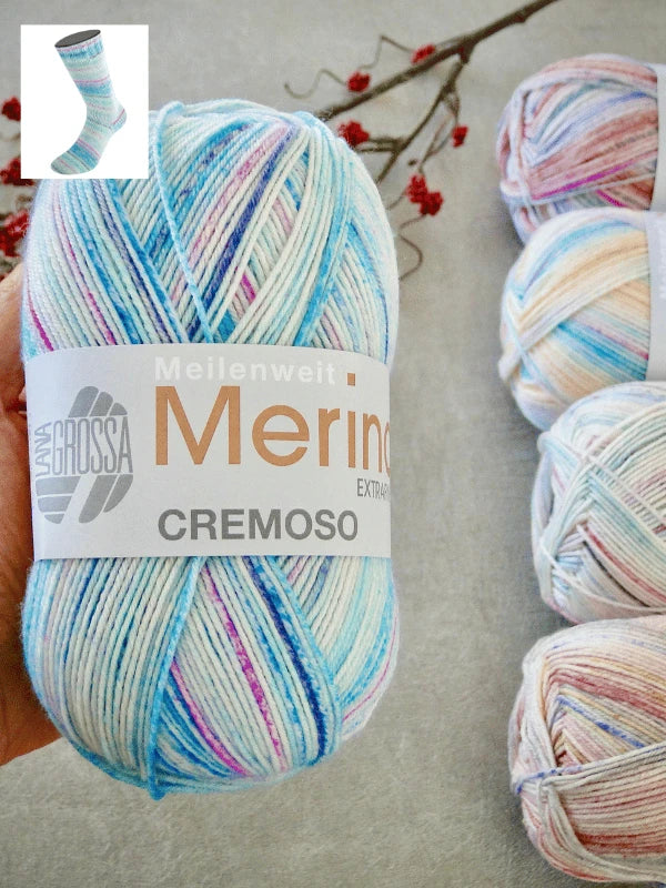 A hand holds a skein of konepestävä Lana Grossa Meilenweit 100 Merino Extrafine Cremoso yarn in pastel blue, pink, and purple. Three more skeins and a twig with red berries are in the background. An inset shows a pastel knit sock.