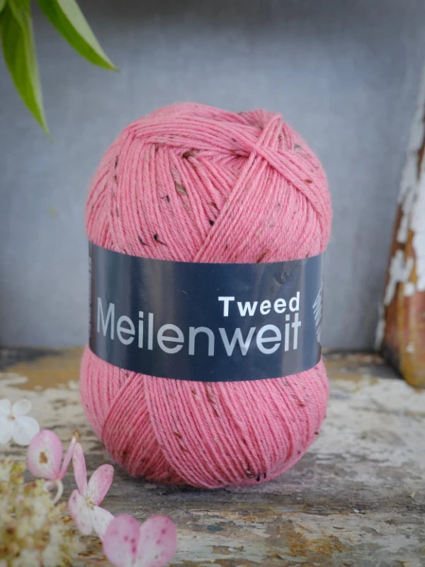 A ball of Lana Grossa Meilenweit 100 Tweed yarn in pink rests on a rustic surface, accompanied by delicate white flowers in one corner and a leafy plant overhead, highlighting the beauty of quality sukkalanka.