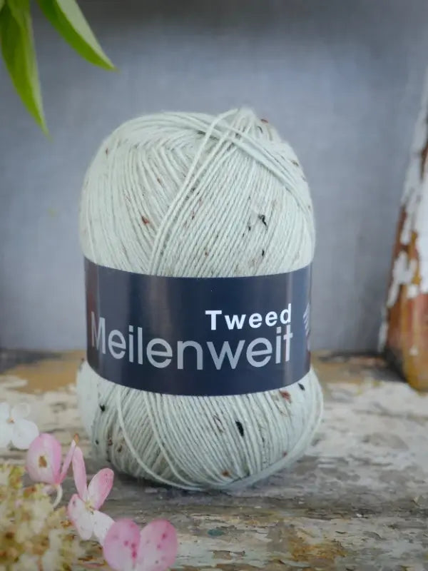 A skein of Lana Grossa Meilenweit 100 Tweed yarn in light grey with subtle dark flecks rests on a rustic wooden surface beside delicate pink and white flowers. This soft sukkalanka brings a charming accent to any knitting project.