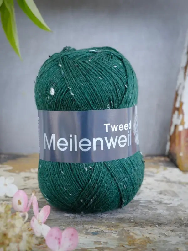 A skein of Lana Grossa Meilenweit 100 Tweed yarn in a deep green tweed color rests on a rustic surface, with blurred greenery and pink flowers in the background—an ideal choice for your next sukkalanka project.