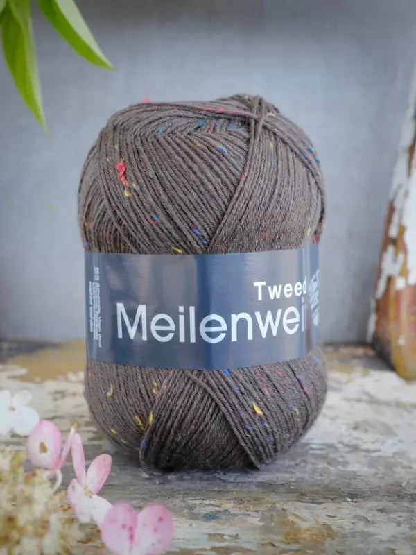 A ball of brown yarn with colorful flecks labeled Lana Grossa Meilenweit 100 Tweed rests on a rustic surface, surrounded by small pink and white flowers. This Lana Grossa Meilenweit 100 Tweed yarn adds charm and texture to any knitting project.